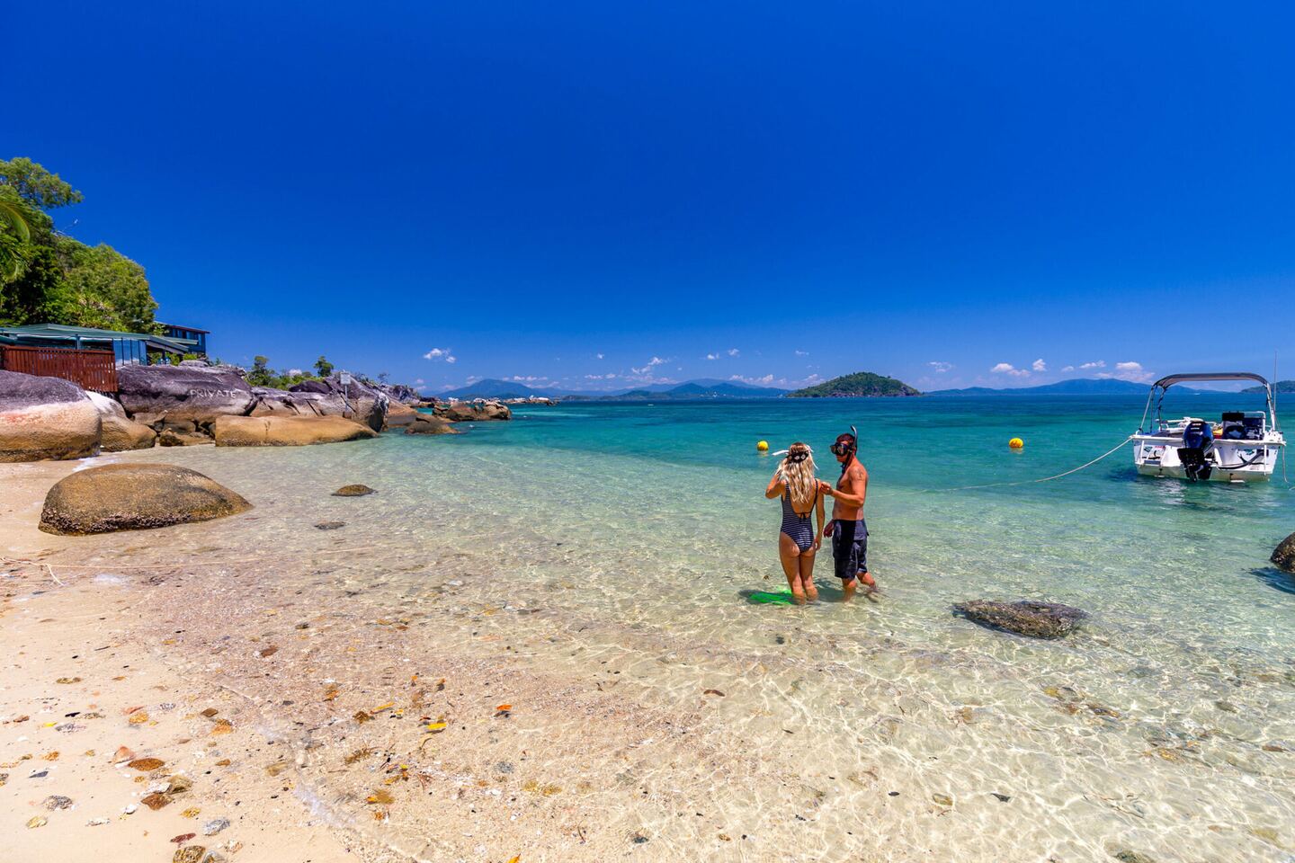 Guests standing in the shallow turquoise water with a boat moored offshore
